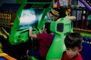Unrecognizable child having fun with bright car racing simulator while turning steering wheel near serious ethnic woman and funny brother in shopping center near playground